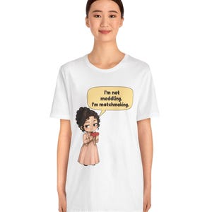 K&ouml;nnte beinhalten: Ein wei&szlig;es T-Shirt mit einer Grafik einer Zeichentrickfigur mit dunklem lockigem Haar, die ein hellrosa Kleid und eine Jacke tr&auml;gt und rote Herzen h&auml;lt. Eine gelbe Sprechblase enth&auml;lt den Text: I'm not meddling. I'm matchmaking.