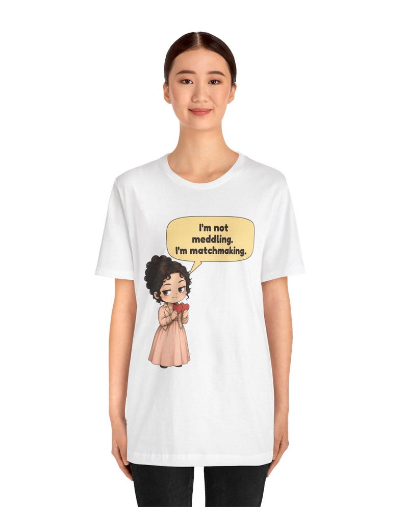 K&ouml;nnte beinhalten: Ein wei&szlig;es T-Shirt mit einer Grafik einer Zeichentrickfigur mit dunklem lockigem Haar, die ein hellrosa Kleid und eine Jacke tr&auml;gt und rote Herzen h&auml;lt. Eine gelbe Sprechblase enth&auml;lt den Text: I'm not meddling. I'm matchmaking.
