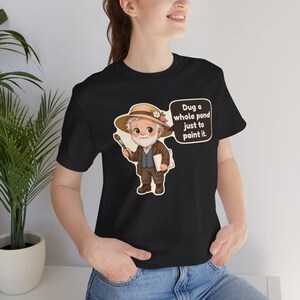 Puede incluir: Una camiseta negra con una ilustraci&oacute;n de dibujos animados de un artista mayor con barba blanca, que lleva un sombrero beige con flores y sostiene un pincel y un lienzo. Un bocadillo contiene el texto: Dug a whole pond just to paint it.