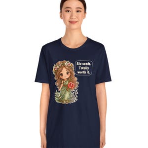 May include: A navy blue short-sleeve t-shirt featuring a graphic of a cartoon character with long brown hair, a flower crown, and a green dress holding a pomegranate. A speech bubble next to the character contains the text: Six seeds. Totally worth it.