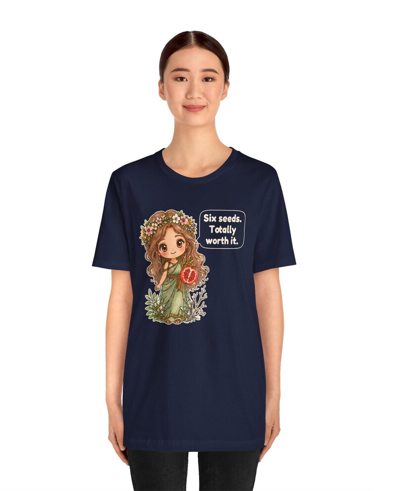 May include: A navy blue short-sleeve t-shirt featuring a graphic of a cartoon character with long brown hair, a flower crown, and a green dress holding a pomegranate. A speech bubble next to the character contains the text: Six seeds. Totally worth it.