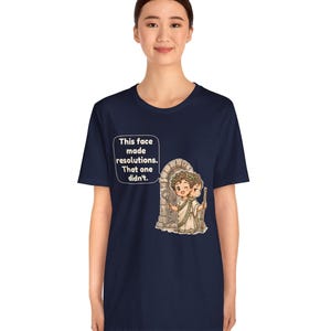K&ouml;nnte beinhalten: Ein marineblaues T-Shirt mit einer Grafik einer r&ouml;mischen Figur mit zwei Gesichtern vor einem Torbogen. Die Figur h&auml;lt einen Schl&uuml;ssel und einen Stab. Eine Sprechblase enth&auml;lt den Text: This face made resolutions. That one didn't.
