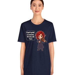 May include: A navy blue t-shirt featuring a graphic of a red-haired character playing a violin. A speech bubble next to the character contains the text: Can't pick a season. Wrote all four. The design is centered on the front of the shirt.