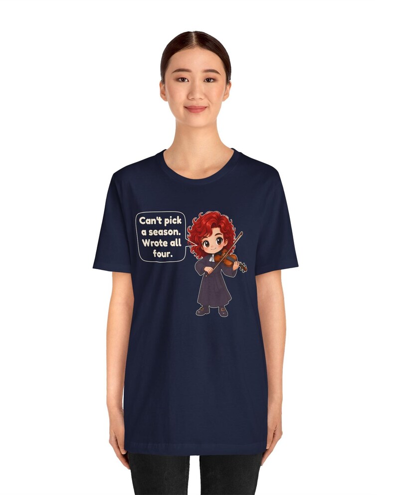 May include: A navy blue t-shirt featuring a graphic of a red-haired character playing a violin. A speech bubble next to the character contains the text: Can't pick a season. Wrote all four. The design is centered on the front of the shirt.