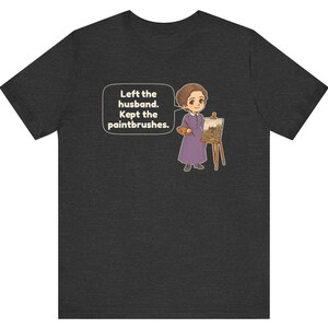 May include: A dark gray t-shirt featuring a cartoon illustration of a person in a purple dress standing next to an easel with a painting. A speech bubble contains the text: Left the husband. Kept the paintbrushes.