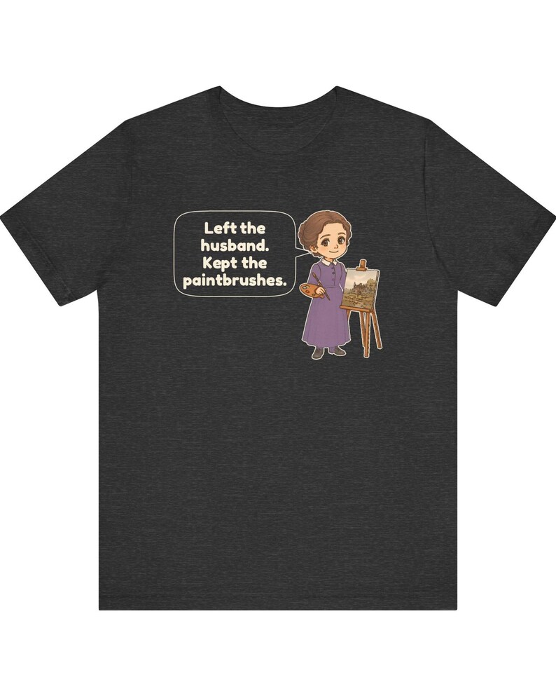 May include: A dark gray t-shirt featuring a cartoon illustration of a person in a purple dress standing next to an easel with a painting. A speech bubble contains the text: Left the husband. Kept the paintbrushes.