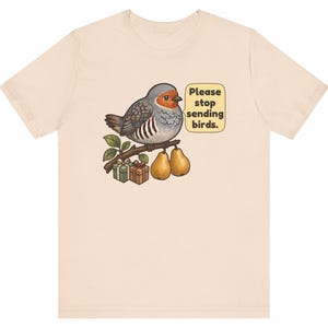 K&ouml;nnte beinhalten: Ein hellbeiges T-Shirt mit einer Grafik eines runden Vogels, der auf einem Ast mit zwei Birnen und zwei kleinen Geschenkkartons sitzt. Eine Sprechblase neben dem Vogel enth&auml;lt den Text, Please stop sending birds. Das Motiv ist mittig platziert.