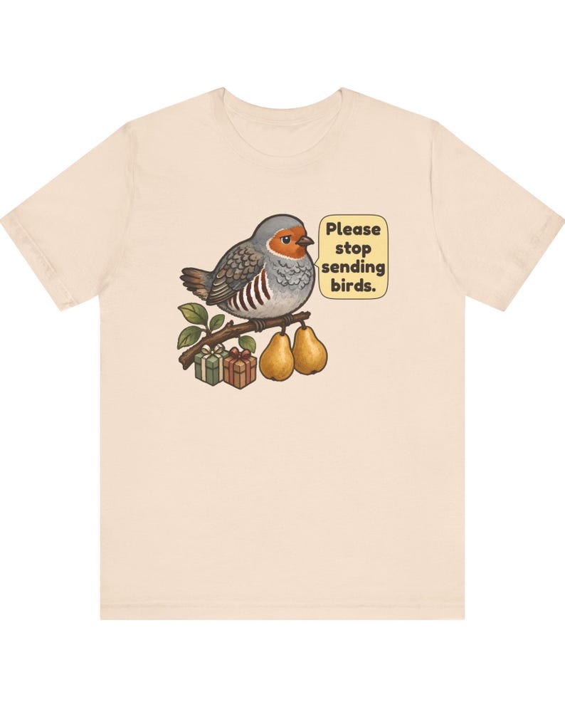 K&ouml;nnte beinhalten: Ein hellbeiges T-Shirt mit einer Grafik eines runden Vogels, der auf einem Ast mit zwei Birnen und zwei kleinen Geschenkkartons sitzt. Eine Sprechblase neben dem Vogel enth&auml;lt den Text, Please stop sending birds. Das Motiv ist mittig platziert.