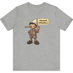 K&ouml;nnte beinhalten: Ein graues T-Shirt mit einer Grafik einer Holzpuppe mit langer Nase, die einen Hut, ein Hemd und eine kurze Hose tr&auml;gt. Eine gelbe Sprechblase neben der Figur enth&auml;lt den schwarzen Text "Honest! (Mostly.)"
