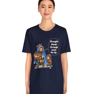 May include: A navy blue short-sleeve t-shirt featuring a graphic of a king in a blue cloak and a child walking through snow. The king carries a basket of food and a speech bubble with the text: Brought dinner through snow. No tip.
