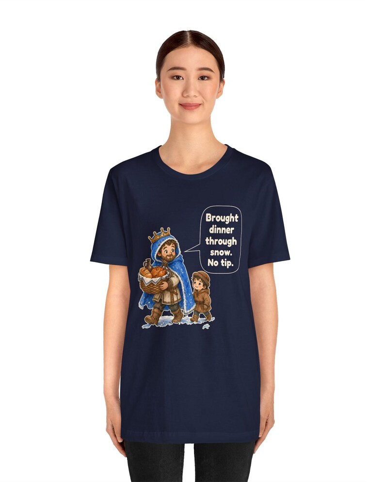 May include: A navy blue short-sleeve t-shirt featuring a graphic of a king in a blue cloak and a child walking through snow. The king carries a basket of food and a speech bubble with the text: Brought dinner through snow. No tip.