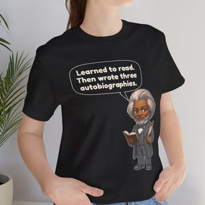 Puede incluir: Una camiseta negra con una ilustraci&oacute;n de dibujos animados de Frederick Douglass sosteniendo un libro y una pluma. Un globo de texto sobre la figura contiene el texto blanco: Learned to read. Then wrote three autobiographies.