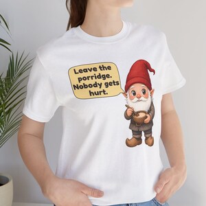 May include: A white t-shirt featuring a graphic of a gnome wearing a red hat and brown clothing, holding a wooden spoon and a bowl of porridge. A speech bubble next to the gnome contains the text: Leave the porridge. Nobody gets hurt.