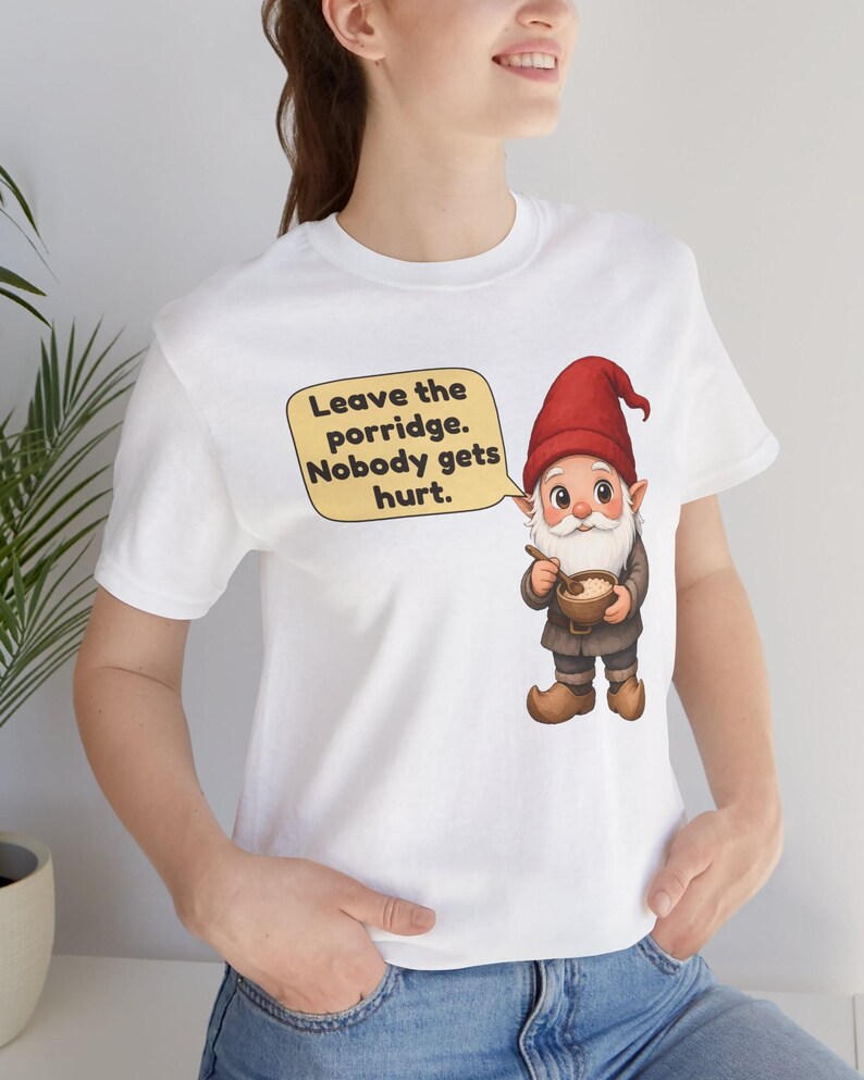 May include: A white t-shirt featuring a graphic of a gnome wearing a red hat and brown clothing, holding a wooden spoon and a bowl of porridge. A speech bubble next to the gnome contains the text: Leave the porridge. Nobody gets hurt.