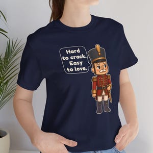 Pu&ograve; includere: Una t-shirt blu navy a maniche corte con la grafica di un soldatino schiaccianoci in uniforme rossa e cappello con finiture dorate. Un fumetto accanto al soldato contiene il testo: Hard to crack. Easy to love.