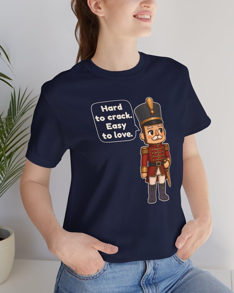 Pu&ograve; includere: Una t-shirt blu navy a maniche corte con la grafica di un soldatino schiaccianoci in uniforme rossa e cappello con finiture dorate. Un fumetto accanto al soldato contiene il testo: Hard to crack. Easy to love.