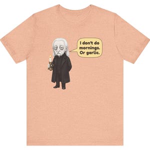 May include: A peach-colored t-shirt featuring a graphic of a pale, long-haired character in a black coat holding a lit candle. A speech bubble next to the character contains the text: I don't do mornings. Or garlic.