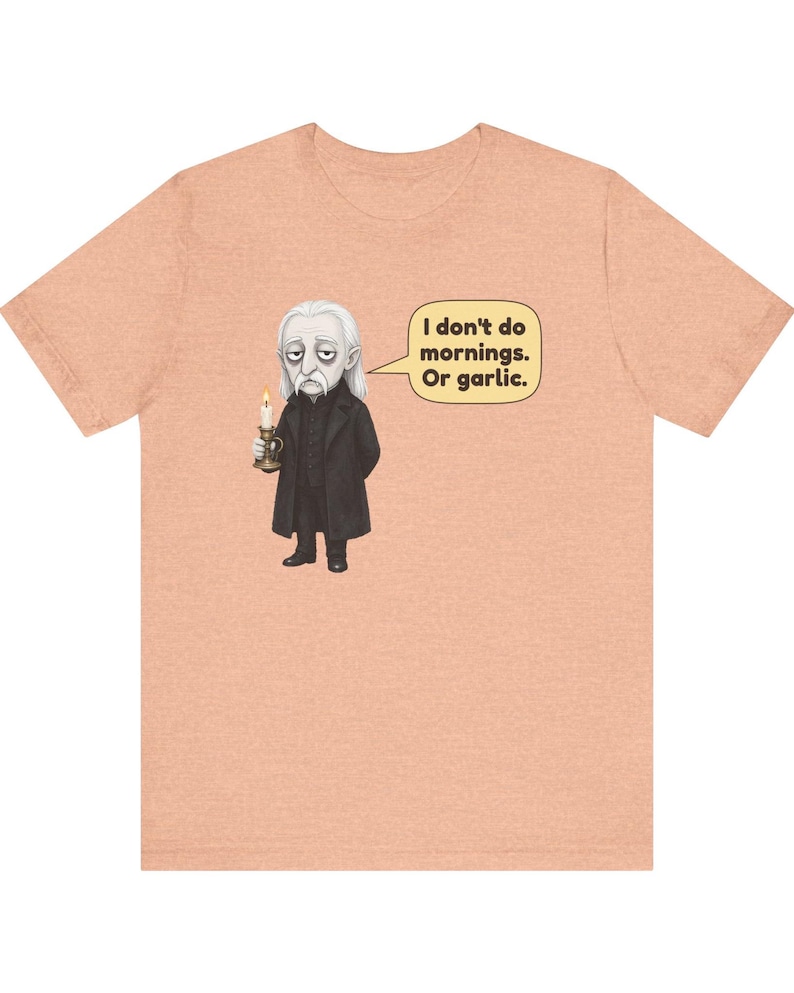 May include: A peach-colored t-shirt featuring a graphic of a pale, long-haired character in a black coat holding a lit candle. A speech bubble next to the character contains the text: I don't do mornings. Or garlic.
