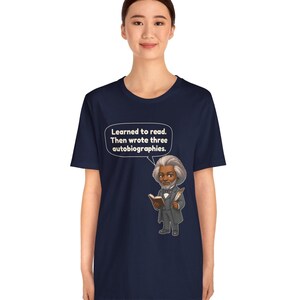 Puede incluir: Una camiseta azul marino de manga corta con una ilustraci&oacute;n de dibujos animados de Frederick Douglass sosteniendo un libro y una pluma. Un globo de texto sobre la figura contiene el texto: Learned to read. Then wrote three autobiographies.