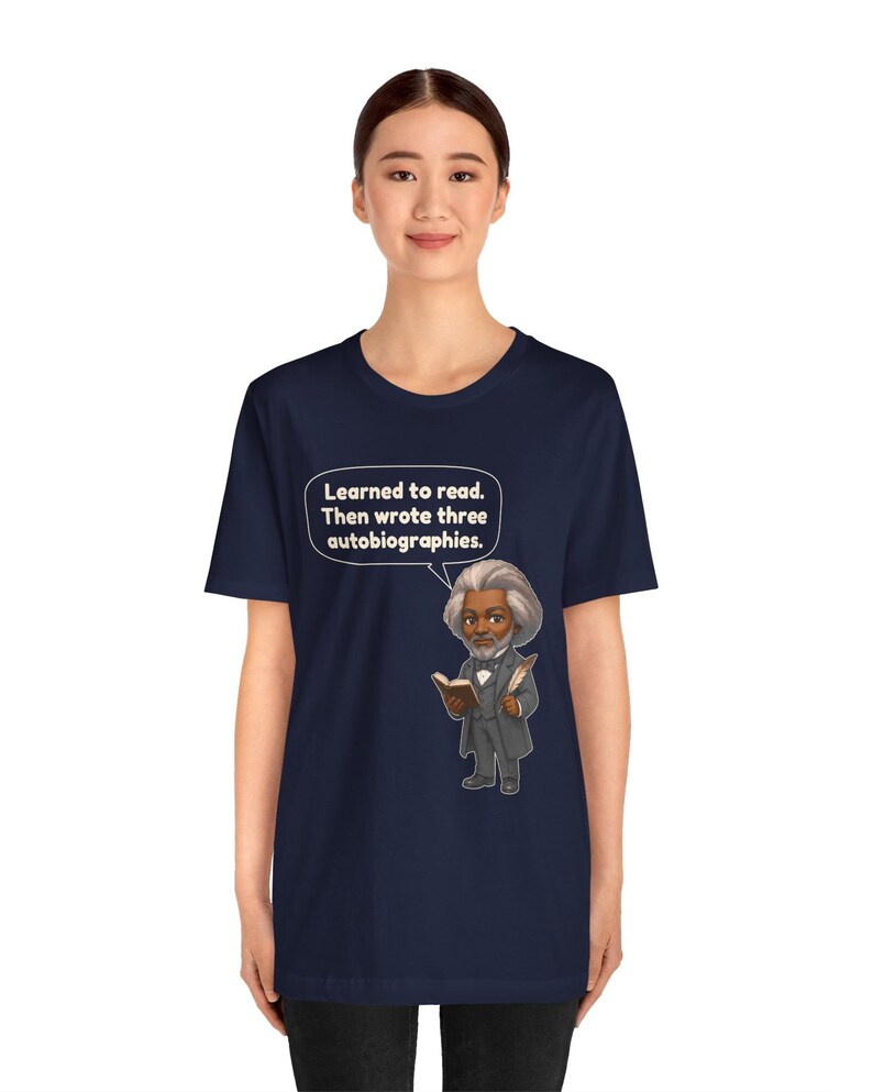 Puede incluir: Una camiseta azul marino de manga corta con una ilustraci&oacute;n de dibujos animados de Frederick Douglass sosteniendo un libro y una pluma. Un globo de texto sobre la figura contiene el texto: Learned to read. Then wrote three autobiographies.