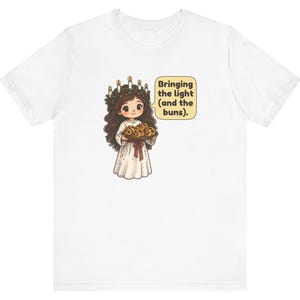 May include: A white t-shirt featuring a graphic of a character with long brown hair wearing a candle crown and a white robe, holding a tray of buns. A speech bubble next to the character contains the text: Bringing the light (and the buns).