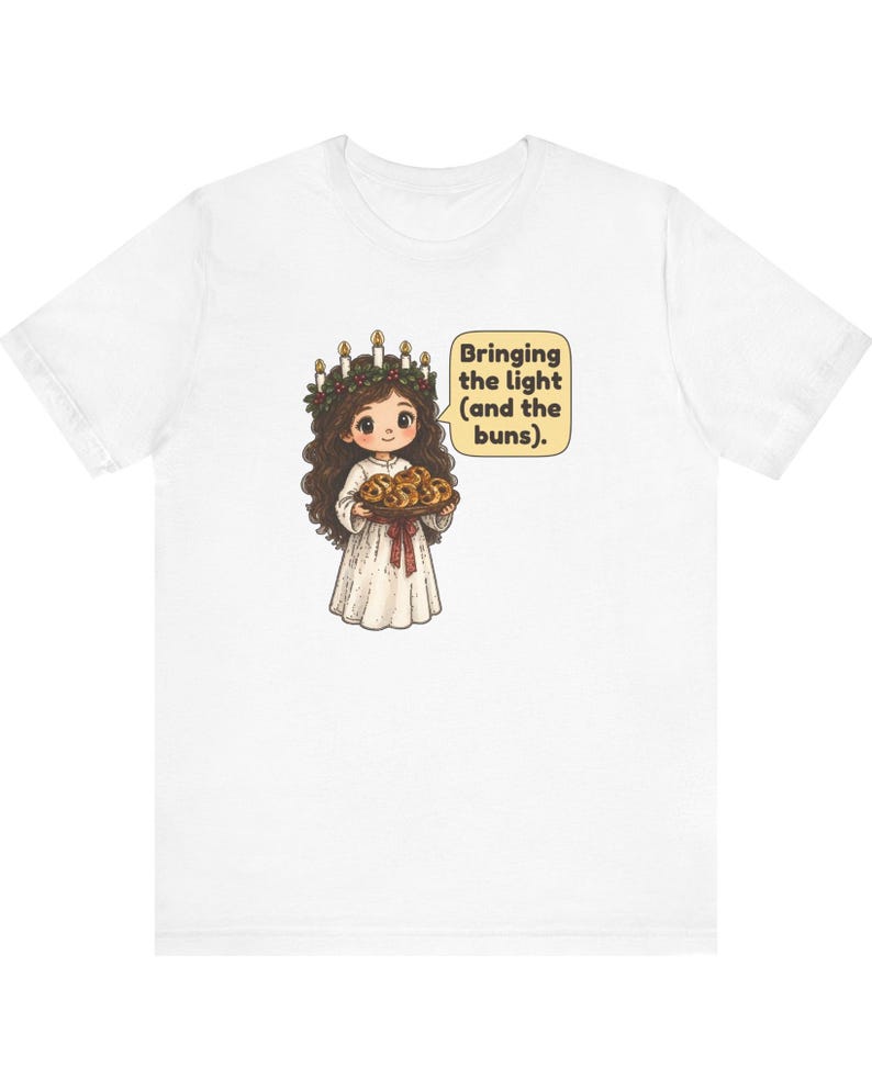 May include: A white t-shirt featuring a graphic of a character with long brown hair wearing a candle crown and a white robe, holding a tray of buns. A speech bubble next to the character contains the text: Bringing the light (and the buns).