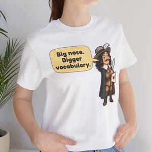 May include: A white short-sleeved t-shirt featuring a cartoon illustration of a historical figure with a large nose, wearing a hat with a feather and a cape. A yellow speech bubble contains the text: Big nose. Bigger vocabulary.