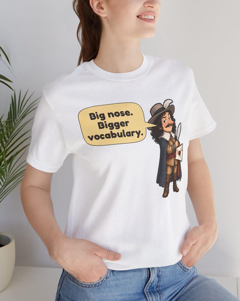 May include: A white short-sleeved t-shirt featuring a cartoon illustration of a historical figure with a large nose, wearing a hat with a feather and a cape. A yellow speech bubble contains the text: Big nose. Bigger vocabulary.