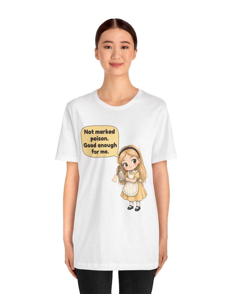 May include: A white short-sleeve t-shirt featuring a cartoon illustration of a girl with blonde hair wearing a yellow dress and white apron. She holds a small glass bottle. A speech bubble above her reads: Not marked poison. Good enough for me.