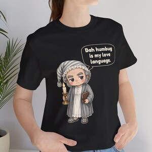 Puede incluir: Una camiseta negra con una ilustraci&oacute;n de dibujos animados de un personaje con gorro de dormir y bata que sostiene una vela y un libro. Un bocadillo sobre el personaje contiene el texto, Bah humbug is my love language. El dise&ntilde;o est&aacute; centrado.