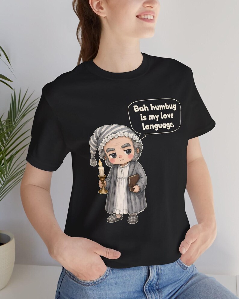 Puede incluir: Una camiseta negra con una ilustraci&oacute;n de dibujos animados de un personaje con gorro de dormir y bata que sostiene una vela y un libro. Un bocadillo sobre el personaje contiene el texto, Bah humbug is my love language. El dise&ntilde;o est&aacute; centrado.