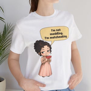 K&ouml;nnte beinhalten: Ein wei&szlig;es T-Shirt mit einer Cartoon-Illustration einer Person mit dunklen lockigen Haaren, die eine beige Jacke und ein rosa Kleid tr&auml;gt. Die Figur h&auml;lt zwei rote Herzen und hat eine gelbe Sprechblase mit dem Text, I'm not meddling. I'm matchmaking.