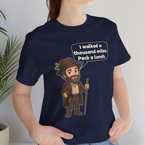 May include: A navy blue t-shirt featuring a cartoon illustration of a bearded hiker wearing a brown hat with a pine sprig, a brown coat, and boots, holding a wooden walking stick. A speech bubble contains the text: I walked a thousand miles. Pack a lunch.