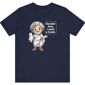 May include: A navy blue t-shirt featuring a cartoon illustration of a bearded figure with curly gray hair wearing a white toga and brown sandals. The figure holds a white towel and a speech bubble reads, Eureka! Also, I need a towel.