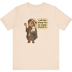 May include: A light beige t-shirt featuring a cartoon illustration of a shepherd holding a small white lamb and a wooden staff. A speech bubble next to the character contains the text: Left the sheep for an angel. Worth it.