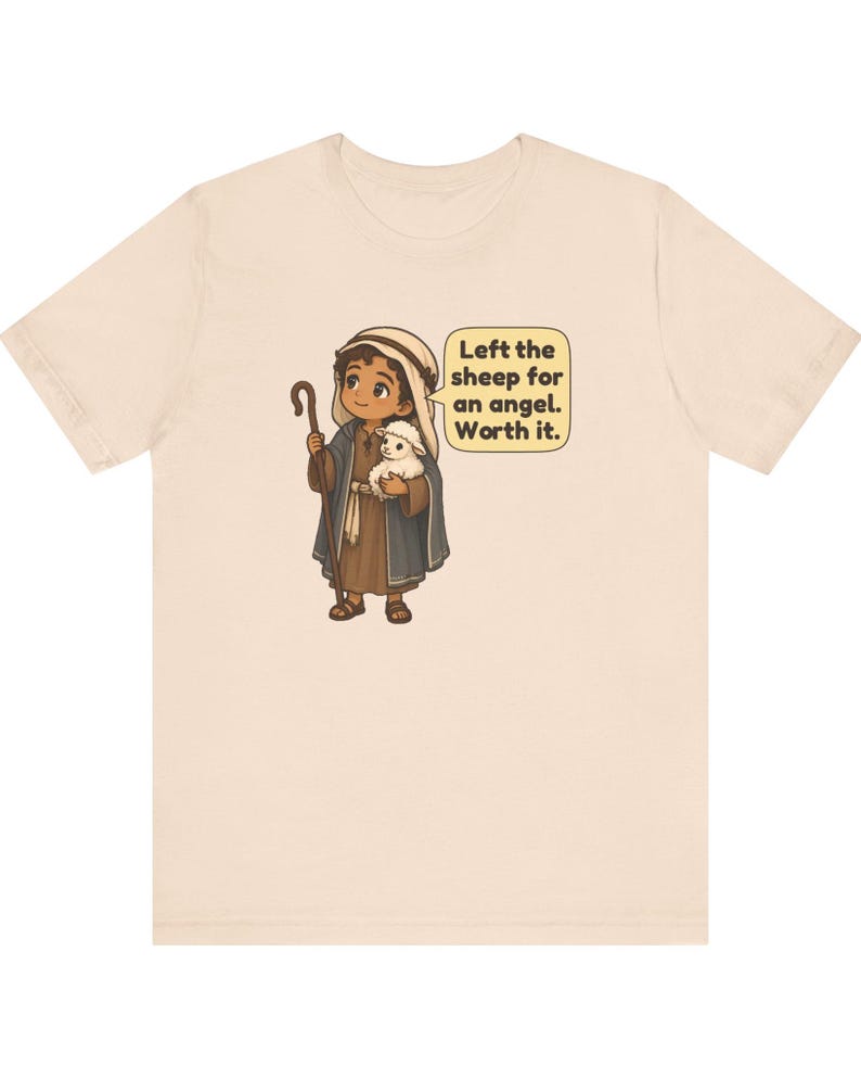 May include: A light beige t-shirt featuring a cartoon illustration of a shepherd holding a small white lamb and a wooden staff. A speech bubble next to the character contains the text: Left the sheep for an angel. Worth it.