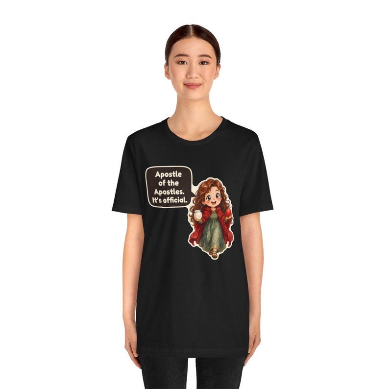May include: A black t-shirt featuring a graphic of a cartoon figure with long brown hair, wearing a red cloak over a green dress, holding a white jar and a red object. A speech bubble contains the text: Apostle of the Apostles. It's official.