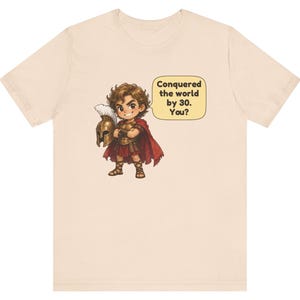 May include: A light beige t-shirt featuring a cartoon illustration of a warrior in bronze armor and a red cape, holding a plumed helmet. A speech bubble next to the character contains the text: Conquered the world by 30. You?