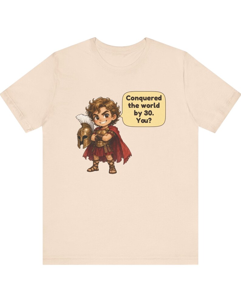 May include: A light beige t-shirt featuring a cartoon illustration of a warrior in bronze armor and a red cape, holding a plumed helmet. A speech bubble next to the character contains the text: Conquered the world by 30. You?
