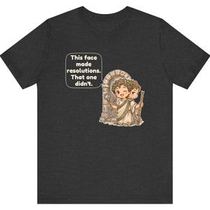 K&ouml;nnte beinhalten: Ein dunkelgraues T-Shirt mit einer Grafik einer r&ouml;mischen Figur mit zwei Gesichtern, die einen Lorbeerkranz und eine Toga tr&auml;gt. Die Figur h&auml;lt einen Schl&uuml;ssel und einen Stab vor einem Steinbogen. Eine Sprechblase enth&auml;lt den Text: This face made resolutions. That one didn't.
