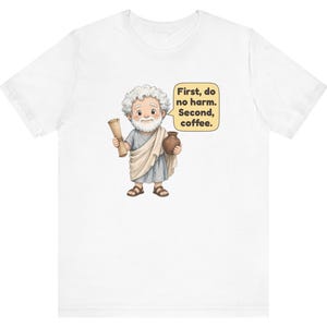 May include: A white t-shirt featuring a cartoon illustration of a bearded man in a toga holding a scroll and a brown pot. A yellow speech bubble next to the figure contains the black text: First, do no harm. Second, coffee.
