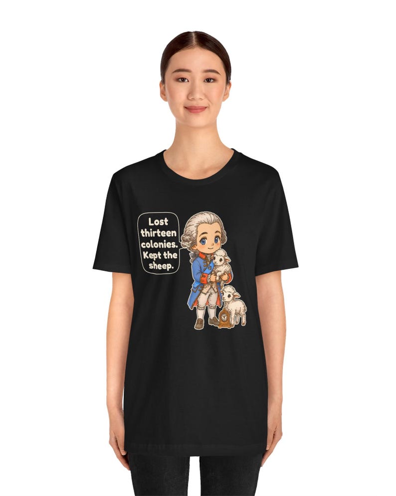 Peut inclure: Un t-shirt noir &agrave; manches courtes avec une illustration de style dessin anim&eacute; d'un personnage historique en manteau bleu tenant un mouton blanc, avec un autre mouton &agrave; c&ocirc;t&eacute;. Le texte indique : Lost thirteen colonies. Kept the sheep.