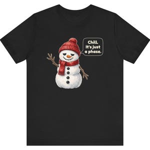 Pode incluir: Uma t-shirt preta de manga curta com o gr&aacute;fico de um boneco de neve a usar um gorro e um cachecol de malha vermelhos. O boneco de neve tem bra&ccedil;os de ramo, um nariz de cenoura e um bal&atilde;o com o texto branco: Chill. It's just a phase.