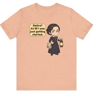 May include: A peach-colored t-shirt featuring a cartoon illustration of a person in a dark dress carrying a satchel and a lit lantern. A speech bubble next to the character contains the text: Retire? At 59 I was just getting started.