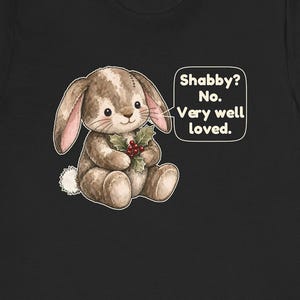 May include: A black t-shirt featuring a graphic of a brown plush bunny holding a sprig of holly with red berries. A speech bubble next to the bunny contains the text: Shabby? No. Very well loved. The bunny has stitched details and a white tail.