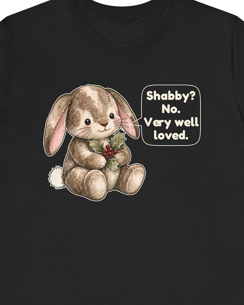 May include: A black t-shirt featuring a graphic of a brown plush bunny holding a sprig of holly with red berries. A speech bubble next to the bunny contains the text: Shabby? No. Very well loved. The bunny has stitched details and a white tail.