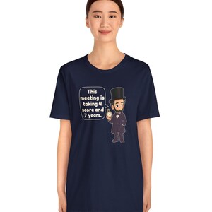 May include: A navy blue t-shirt featuring a cartoon illustration of Abraham Lincoln wearing a top hat and holding a pocket watch. A speech bubble next to the figure contains the text: This meeting is taking 4 score and 7 years.