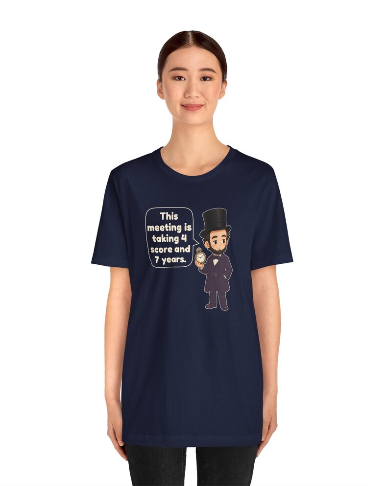 May include: A navy blue t-shirt featuring a cartoon illustration of Abraham Lincoln wearing a top hat and holding a pocket watch. A speech bubble next to the figure contains the text: This meeting is taking 4 score and 7 years.