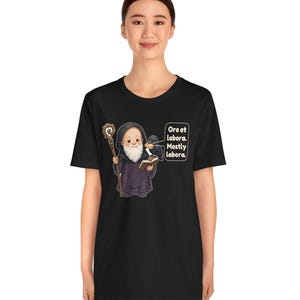 May include: A black t-shirt featuring a cartoon monk with a white beard, holding a wooden staff and an open book. A black raven sits on the monk's shoulder. A speech bubble contains the text: Ora et labora. Mostly labora.
