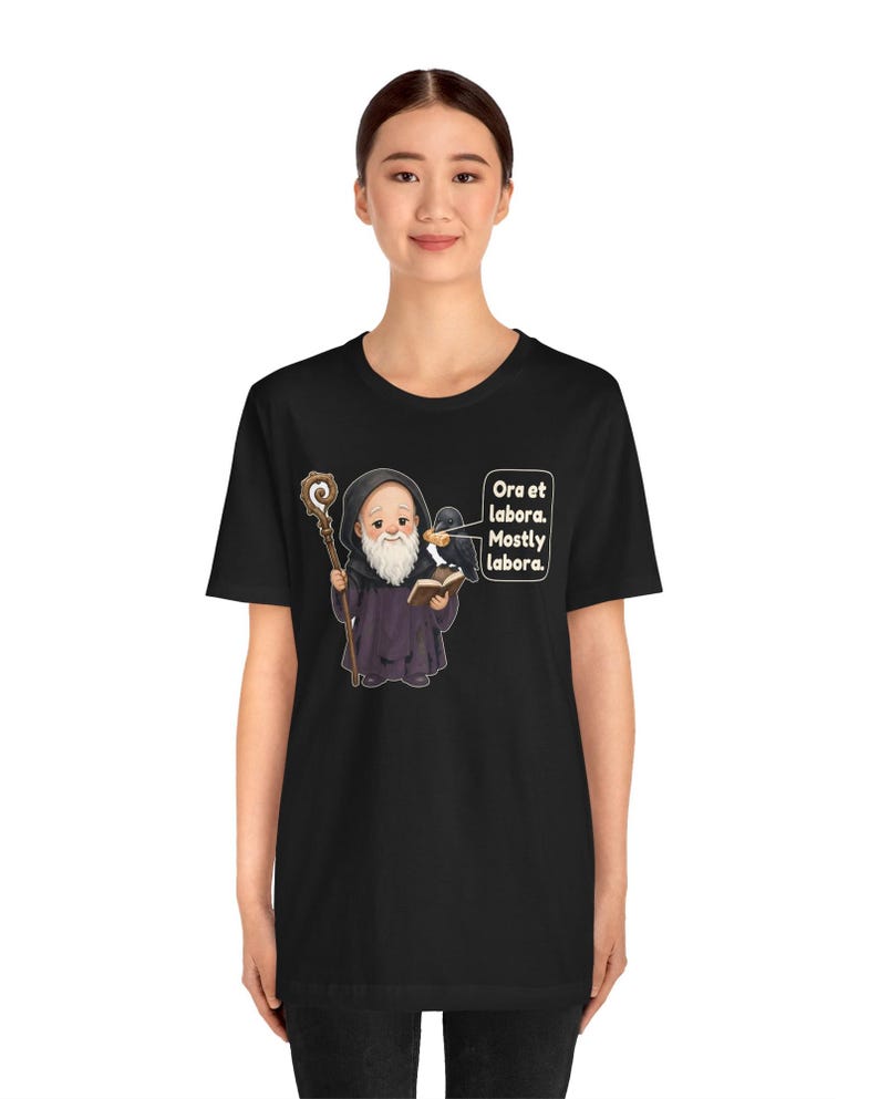 May include: A black t-shirt featuring a cartoon monk with a white beard, holding a wooden staff and an open book. A black raven sits on the monk's shoulder. A speech bubble contains the text: Ora et labora. Mostly labora.